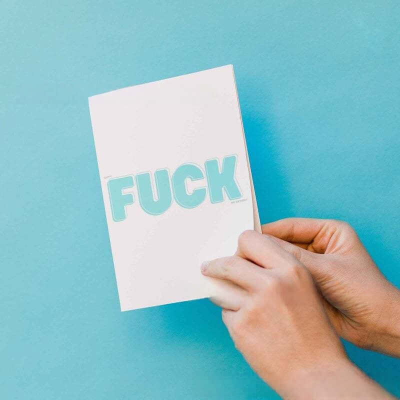 Hand holding a humorous birthday card featuring bold lettering of "F*CK" against a bright blue background.