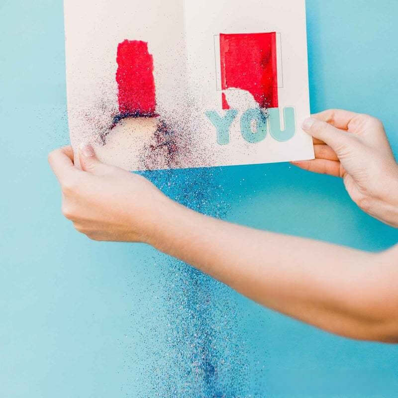 Hands revealing a colorful glitter bomb card titled 'YOU', creating a playful surprise and laughter for a birthday.
